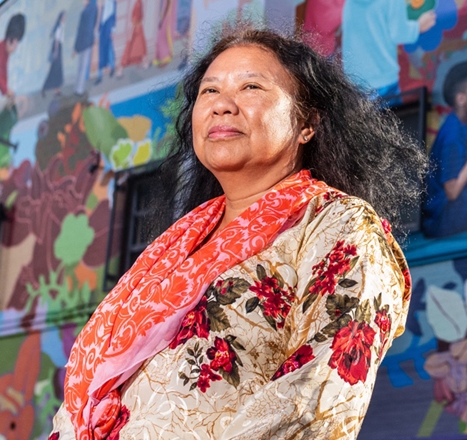 Rashidah Salam wears a brightly colored scarf over a patterned shirt while standing in front of her mural.